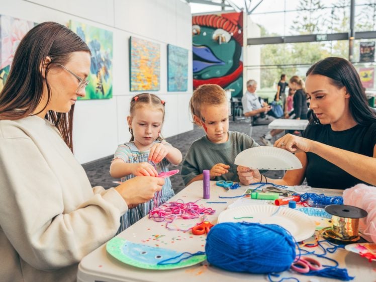 Image of Two women and two young children sit at a table covered with craft supplies, including wool, paper plates, and scissors, making art projects together in a bright room with colourful artwork on the walls.