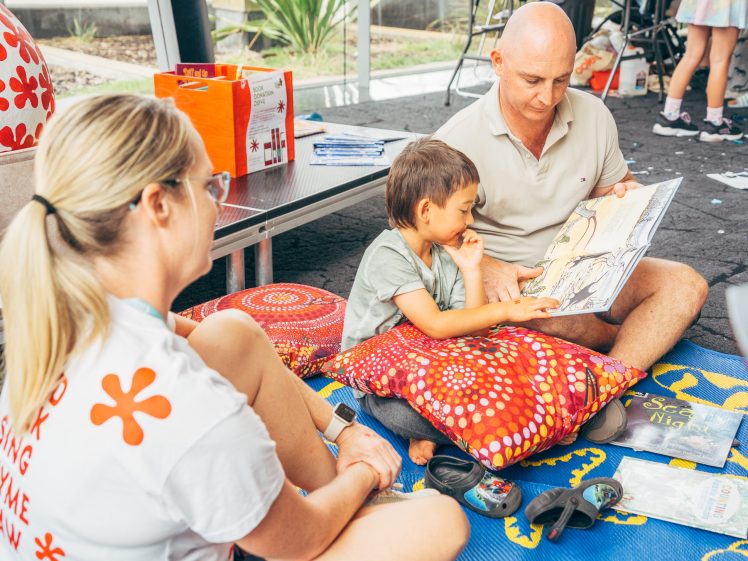 Image of A man sits on the floor reading a picture book to a young boy, whilst a woman sits nearby watching. They are surrounded by colourful cushions and art supplies, in a bright, relaxed indoor setting.