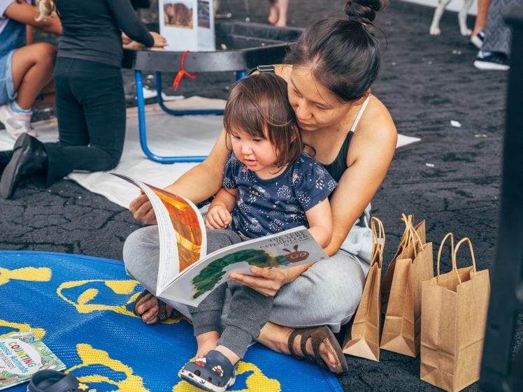 Image of A woman sits on a blue rug with a young child on her lap, reading a picture book together. Paper bags and more books are nearby. Other people are blurred in the background.