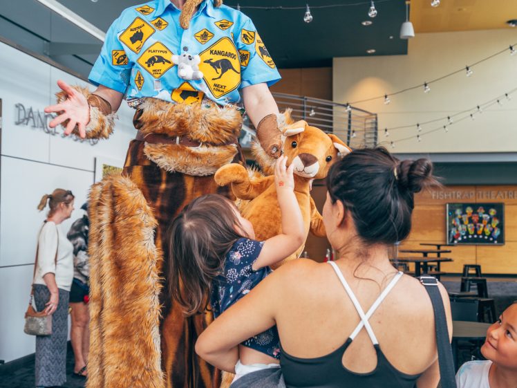 Image of A performer on stilts dressed in a colourful kangaroo costume interacts with a smiling child held by an adult in a busy indoor space, whilst another child looks on.