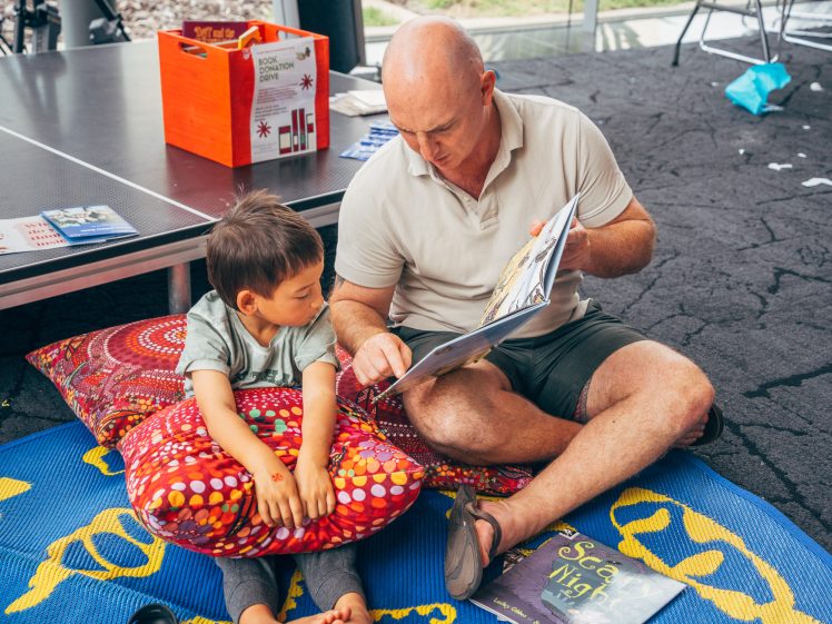 Image of A bald man sits cross-legged on a colourful mat, showing a picture book to a young boy beside him. They are indoors, surrounded by books, cushions, and a box labelled BOOK SWAP.
