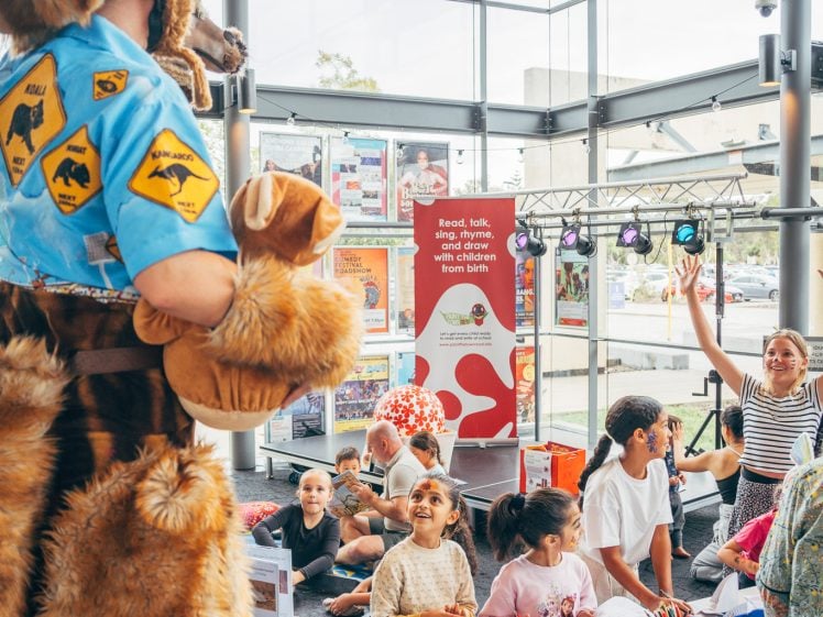 Image of A person in a large animal costume stands near a group of children who are drawing and crafting at tables in a bright, modern indoor space. One child is raising their hand excitedly.