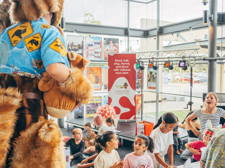 Image of A person in a kangaroo costume interacts with a group of children doing crafts at a table in a brightly lit room. Adults supervise, and the background features large windows and a colourful sign.