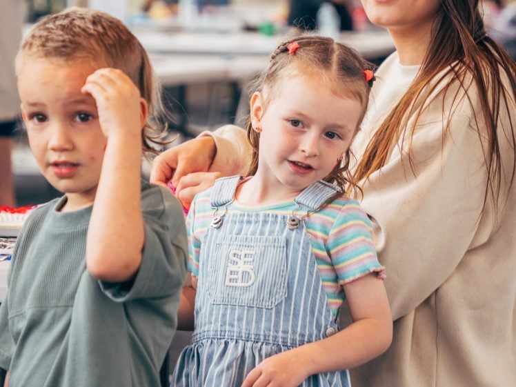 Image of A woman with long brown hair and glasses smiles whilst standing with two young children, a girl in a blue striped dress and a boy in a green shirt, in a bright indoor setting.