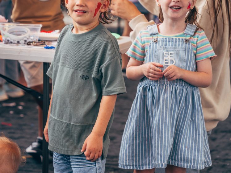 Image of Two young children with face paint smile at the camera at an indoor event. The boy wears a green shirt and shorts, and the girl wears a striped dress. Other children and adults are seen in the background at a craft table.