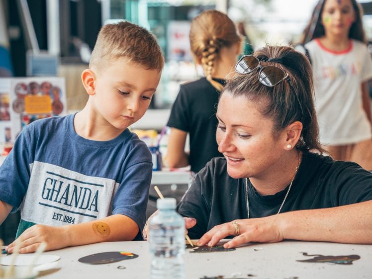 Image of A woman and a young boy sit at a table doing arts and crafts together. The woman is smiling and using a tool whilst the boy watches. Other children are visible in the background. A water bottle sits on the table.