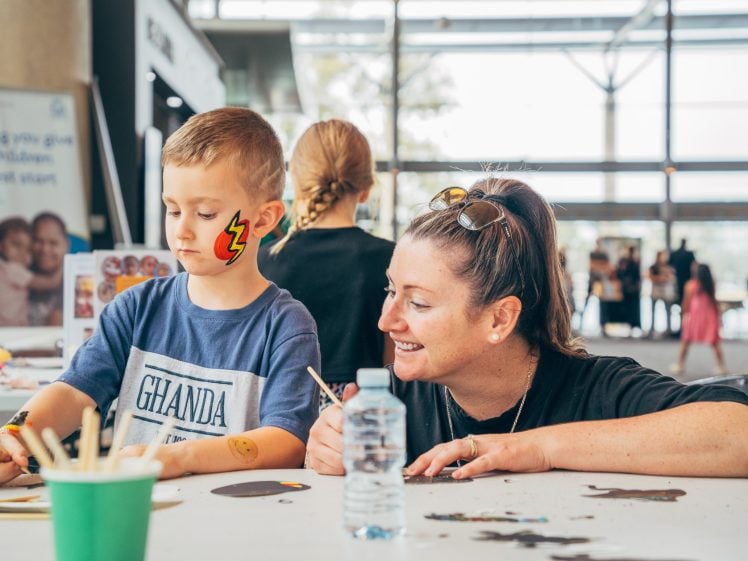 Image of A young boy with a lightning bolt face painting sits at a table painting, whilst a woman beside him smiles and watches. Art supplies and a water bottle are on the table, with a bright, airy background and people in the distance.
