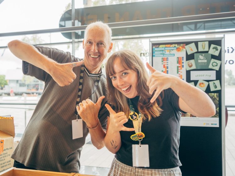 Image of Two people smiling and giving enthusiastic thumbs-up and shaka hand signs at an indoor event, both wearing name badges. A display board with colourful graphics is visible in the background.