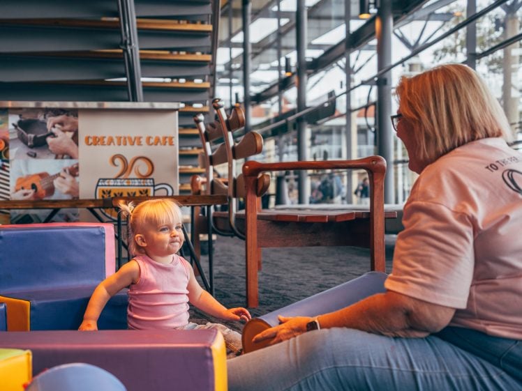 Image of A toddler in a pink outfit smiles whilst playing on soft blocks indoors, as an older woman with blonde hair and glasses sits nearby, smiling back. A “Creative Café” sign is visible in the background.