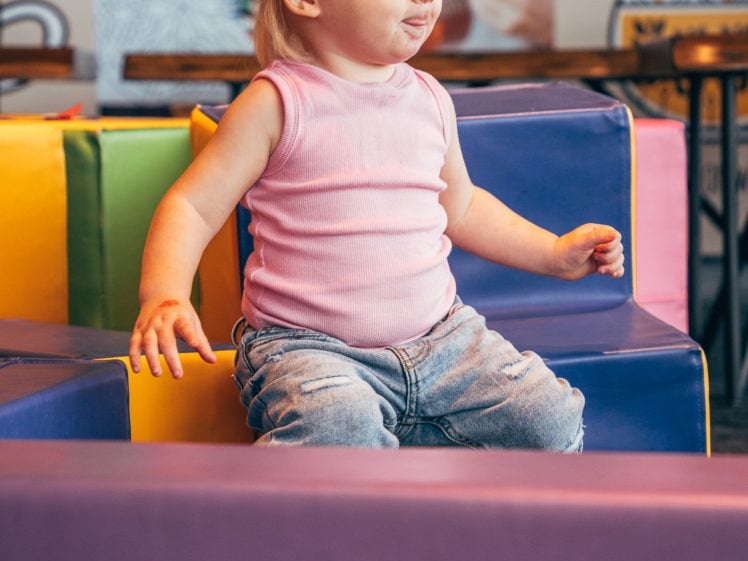 Image of A young child with blonde hair in a pink sleeveless top and jeans sits on colourful foam blocks in a play area, looking to the side with a thoughtful expression.