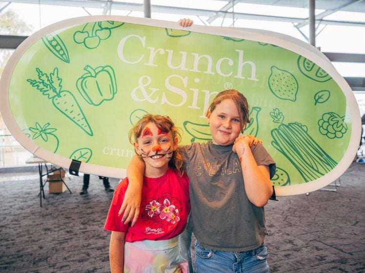 Image of Two smiling children stand together in front of a large green Crunch & Sip sign. One child has face paint resembling a cat and wears a red shirt; the other, in a grey shirt, has an arm round their friend.