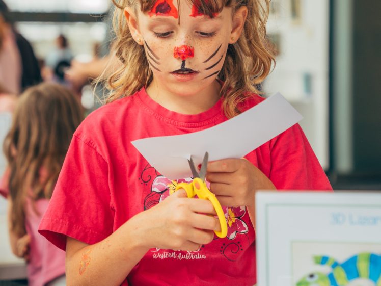 Image of A young girl with cat face paint cuts a piece of paper with yellow scissors at a craft table, focused on her activity. Other children and adults are visible in the background.