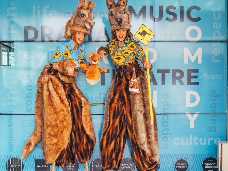 Image of Two performers on stilts wearing kangaroo costumes and holding a toy and a kangaroo crossing sign stand in front of a blue Mandurah Performing Arts Centre wall covered with creative words.
