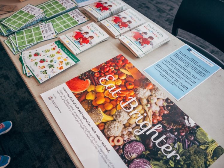 Image of A table displays brochures, pamphlets, sticker sheets with fruit and vegetables, and a large Eat Brighter poster featuring colourful produce, promoting healthy eating and nutrition education.