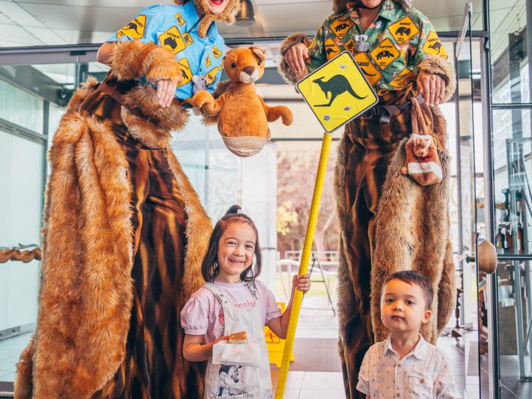Image of Two people on stilts in kangaroo costumes pose indoors with a girl and a boy. The performers hold a toy kangaroo and a yellow kangaroo crossing sign. The children smile, and the scene appears festive and playful.
