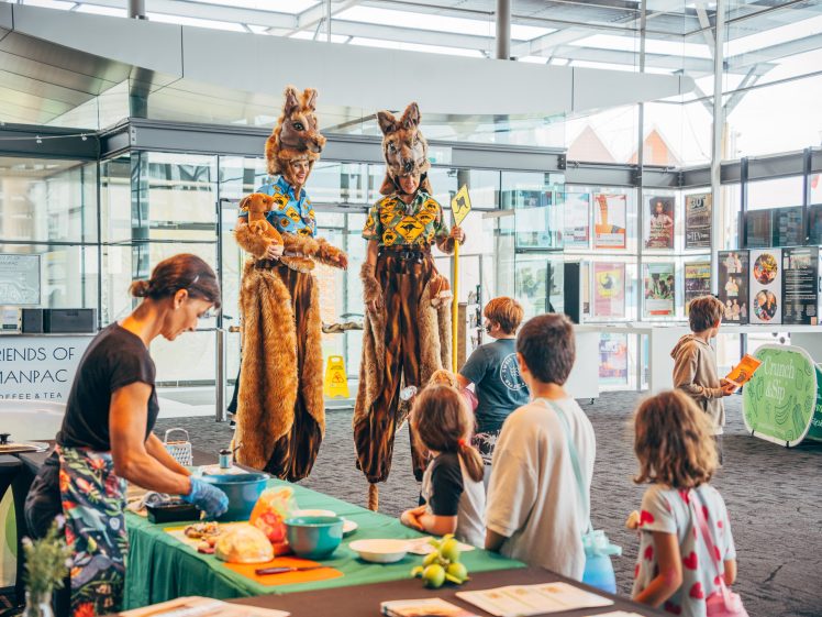 Image of Two people in tall kangaroo costumes entertain children at an indoor event. A woman prepares food samples at a nearby table whilst children watch and interact, surrounded by posters and a bright, modern space.