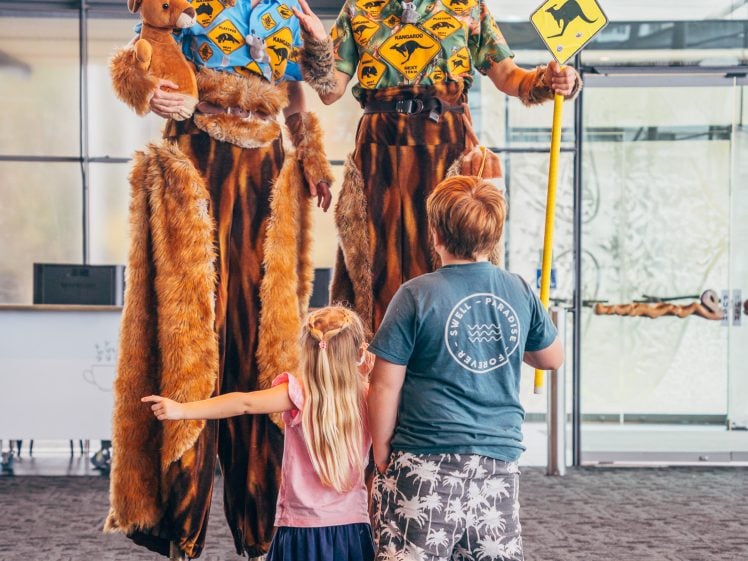 Image of Two adults on stilts dressed as kangaroos and smiling pose with two children; one child hugs the other while holding a kangaroo crossing sign. The adults wear furry costumes and kangaroo headpieces.