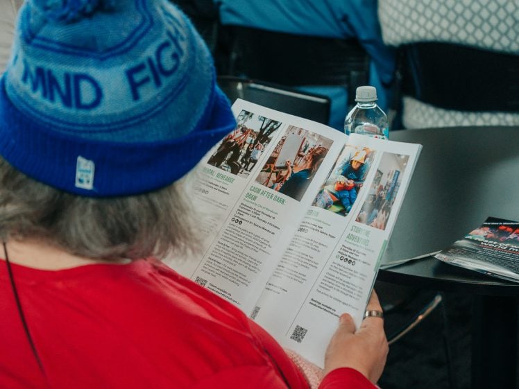 Image of A person in a blue hat and red top sits among a crowd, reading a magazine or programme at a table with a water bottle and another magazine beside them. The focus is on the reader from behind.