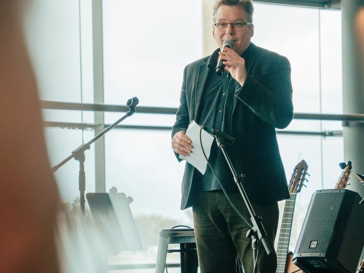 Image of A man in a dark suit stands on a stage holding a microphone and a piece of paper, speaking to an audience. Musical instruments, including guitars and a saxophone, are visible beside him. Large windows are in the background.