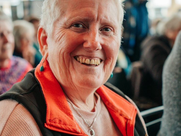 Image of An older woman with short grey hair, wearing a light pink top and a black waistcoat with an orange lining, smiles warmly whilst sitting indoors among a group of people.