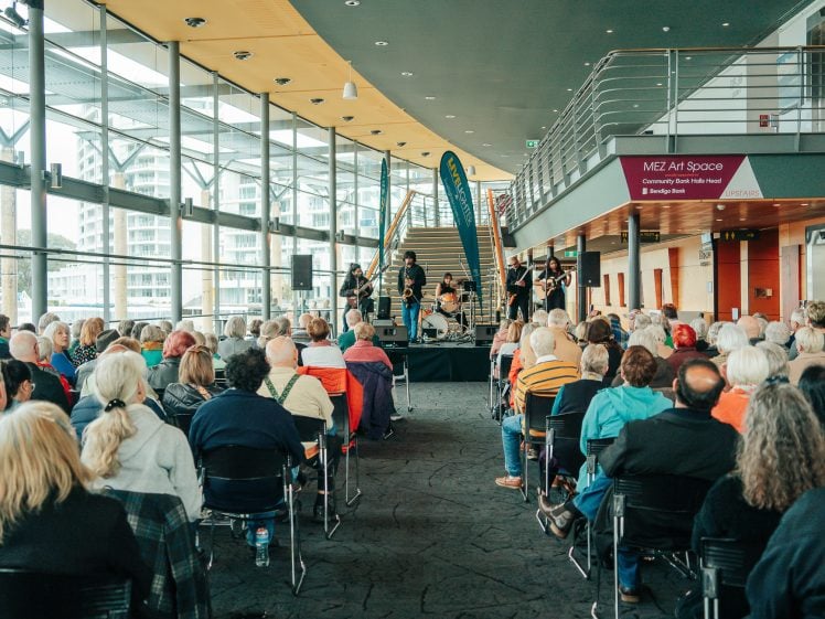 Image of A band performs on stage in a modern indoor venue with large windows, as a seated audience watches attentively. The space is bright, with many people and a sign reading “MET Art Space” on the wall.