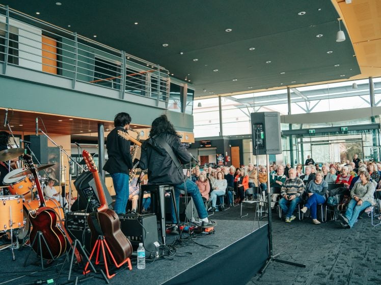 Image of A band performs on stage with guitars and drums in a modern indoor venue, facing an audience seated and watching attentively. Natural light fills the spacious room through large windows.