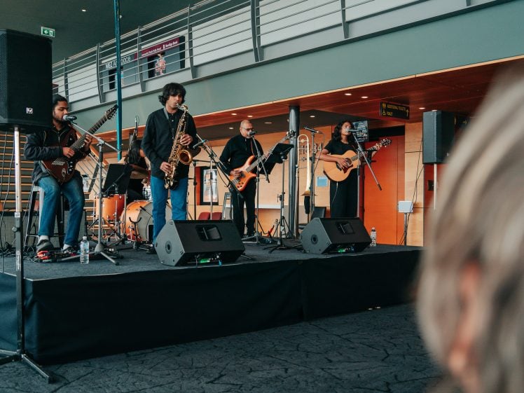 Image of A five-member band performs on an indoor stage with guitars, a saxophone, and microphones. The musicians are standing and playing their instruments. Audience members are partially visible in the foreground.