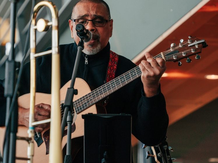 Image of A man wearing glasses and a black jacket plays an acoustic guitar and sings into a microphone. A trombone stands on a nearby stand, and the background features modern architecture with metal and glass elements.