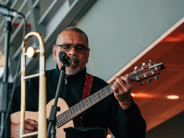 Image of A man wearing glasses plays an acoustic guitar and sings into a microphone on stage. A trombone and another guitar are visible nearby, with modern architectural elements in the background.