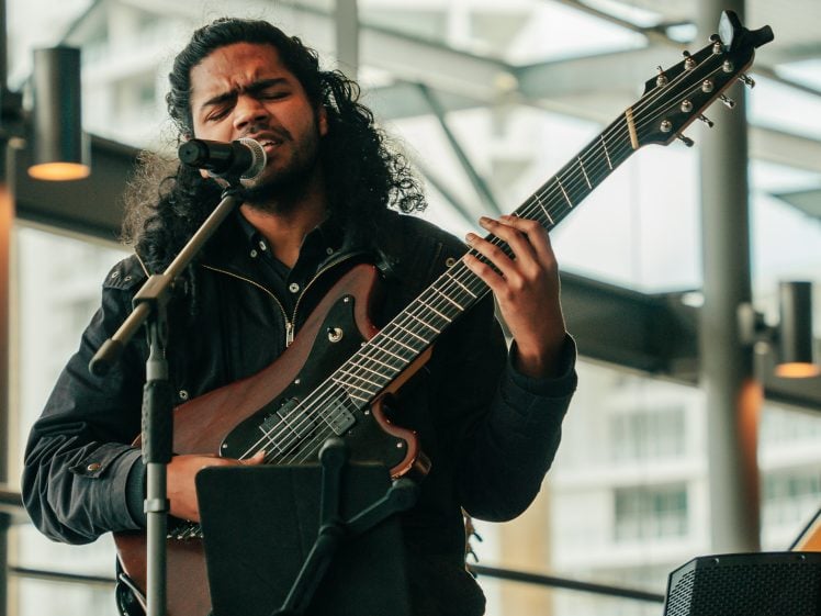 Image of A musician with long curly hair passionately sings into a microphone whilst playing an electric guitar on stage indoors, with modern glass windows and buildings in the background.