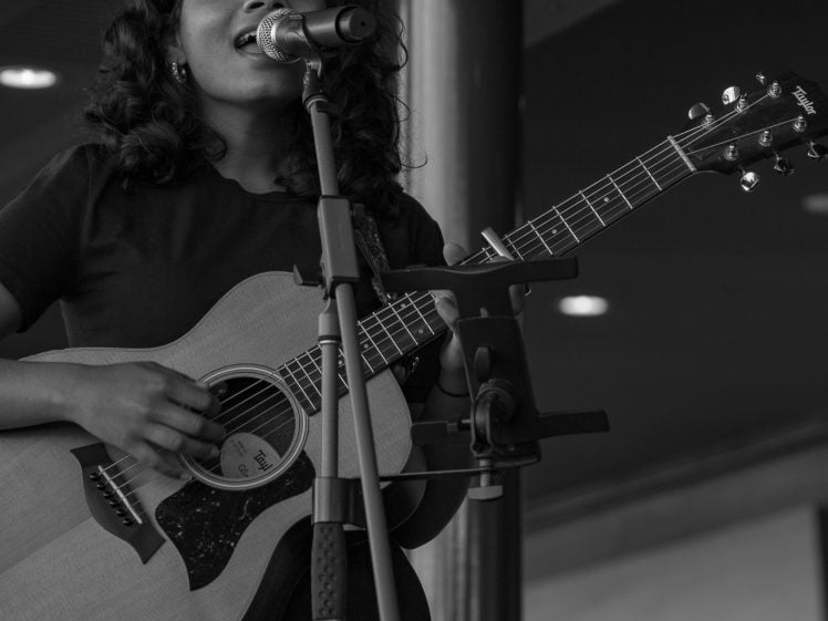 Image of A person with curly hair sings passionately into a microphone whilst playing an acoustic guitar on stage. The image is in black and white.