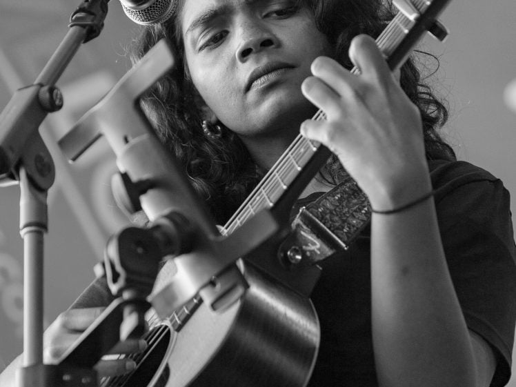 Image of A young woman with curly hair plays an acoustic guitar and sings into a microphone on stage, focused and intent. The photo is in black and white.