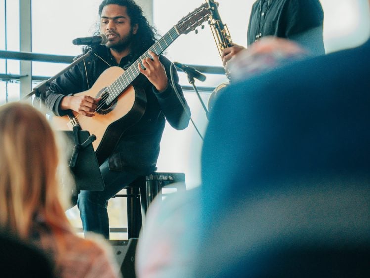 Image of Two musicians perform indoors; one plays an acoustic guitar while seated and the other plays a saxophone standing beside him. They are in front of large windows, with an audience visible in the foreground.