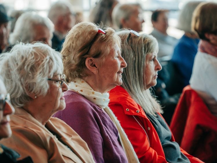 Image of A group of elderly women sit in a row, attentively watching something out of view at an indoor event. The background shows other older adults also seated and engaged.