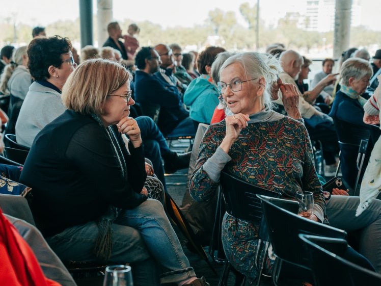 Image of A group of older adults sit together indoors, engaged in conversation. Two women in the foreground talk closely, whilst others listen or chat in the background. The atmosphere seems lively and sociable.