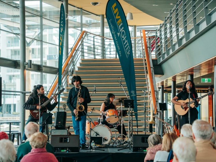 Image of A band performs on an indoor stage in front of an audience. The band includes a guitarist, saxophonist, drummer, and another guitarist, with LIVELIGHTER banners displayed beside a staircase behind them.