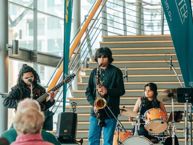 Image of A small band performs indoors near a staircase, featuring a guitarist, saxophonist, and drummer. Audience members watch from seated rows, and LiveLighter banners are displayed behind the band.
