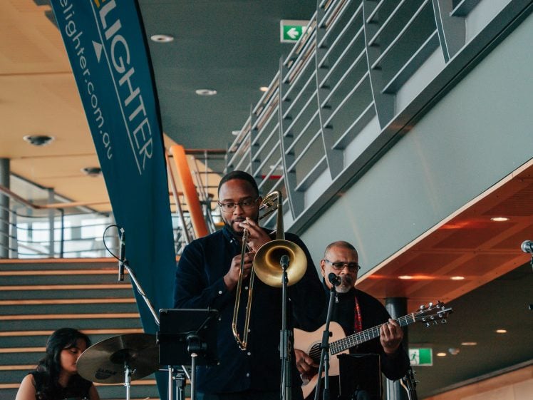 Image of A band performs indoors with a drummer, a trombone player, and a guitarist. A large “LiveLighter” banner stands nearby, and musical equipment is visible on stage. The setting appears modern with metal railings and stairs.