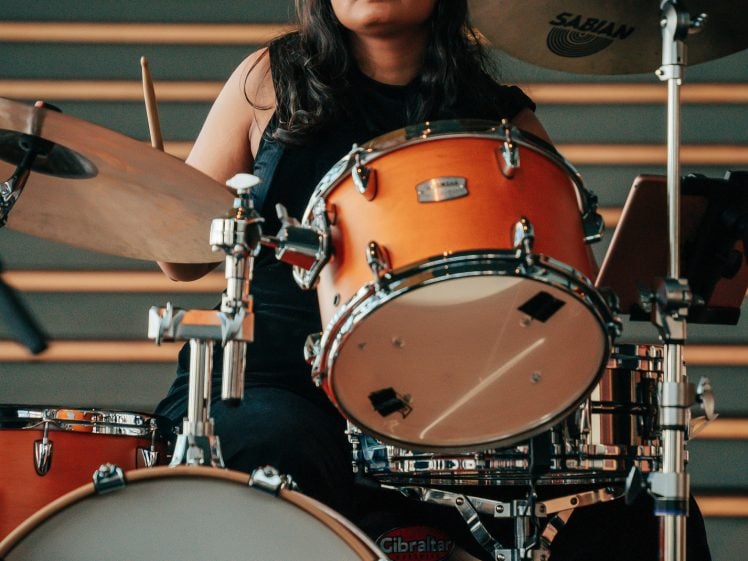 Image of A person with long dark hair plays a drum kit on stage, looking focused. The drum kit includes cymbals and various drums. The background shows horizontal lines, likely steps or panelling.