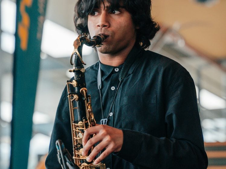 Image of A young person with medium-length dark hair plays a saxophone indoors, wearing a dark button-up shirt. There is a microphone in front of them and a blurred background with stairs and a banner.