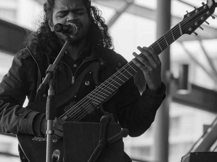 Image of A musician with long curly hair sings into a microphone while playing a six-string bass guitar. He is seated indoors with a modern, industrial-style background. The image is in black and white.