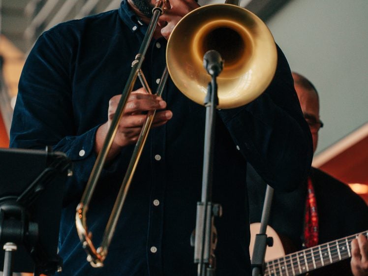 Image of A man wearing glasses and a dark shirt plays a trombone on stage, standing behind a microphone, with another musician and music stands visible in the background.