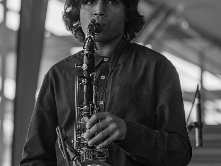 Image of A young person with medium-length hair plays a saxophone indoors, looking at the camera. The scene is black and white, with blurred backgrounds and a microphone stand visible behind them.