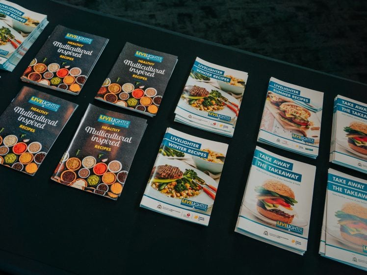 Image of A table displays multiple stacks of healthy recipe booklets, including Multicultural Inspired Recipes, Winter Recipes, and Take Away the Takeaway, each with colourful food images on their covers.