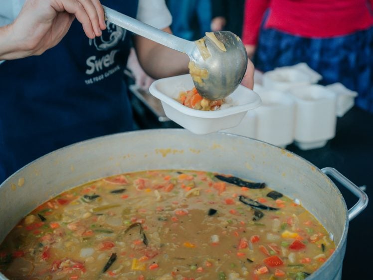 Image of A person ladles vegetable soup from a large pot into a bowl, with several bowls lined up in the background, suggesting a community meal or food service.