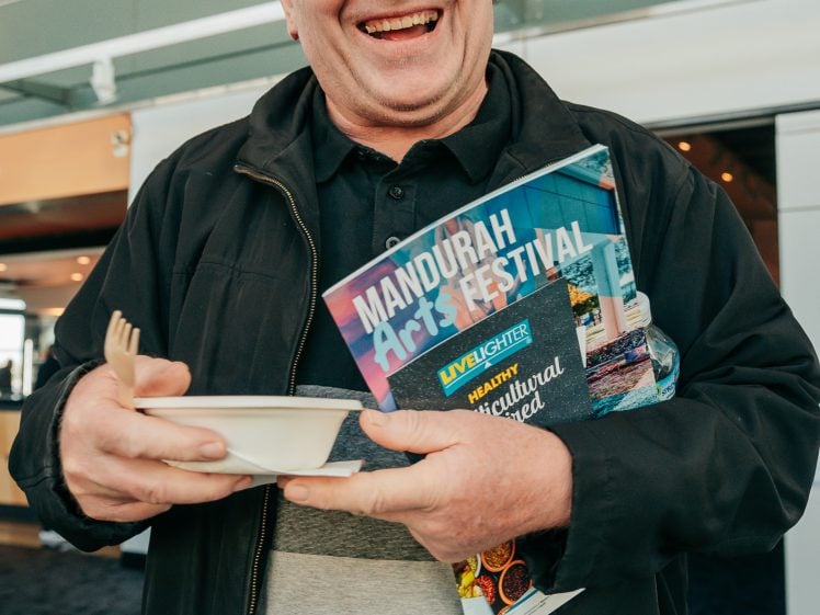 Image of A smiling man wearing glasses and a black jacket holds a bowl with a wooden fork in one hand and Mandurah Arts Festival leaflets in the other, standing indoors.