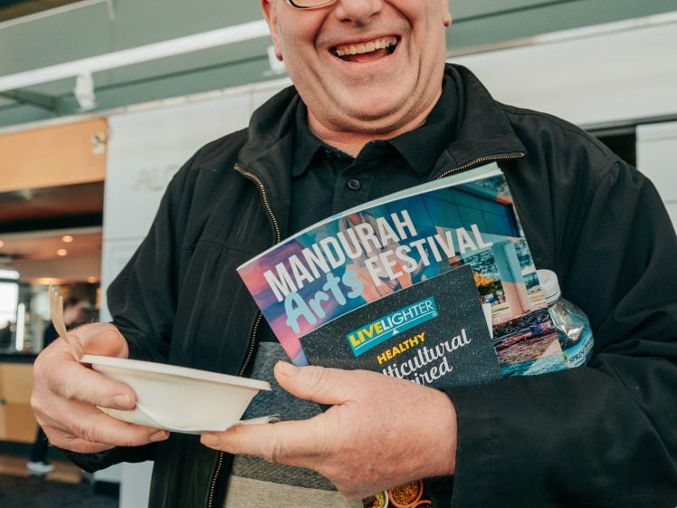 Image of A smiling bald man wearing glasses holds a bowl and several leaflets, including one for the Mandurah Arts Festival, while standing indoors.