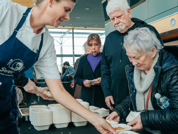 Image of A chef in a blue apron serves food to an elderly woman at a table, whilst other people wait in a queue. The setting appears to be indoors with natural light and stacked food containers on the table.