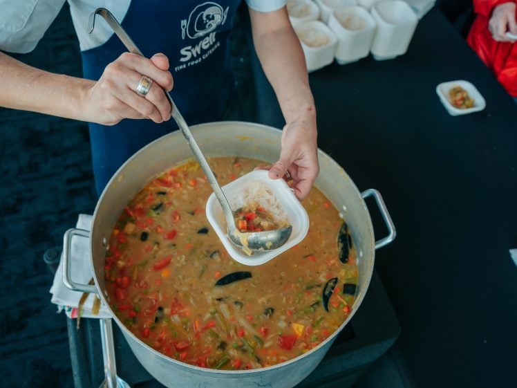 Image of A person in a blue apron ladles soup with vegetables and seafood from a large pot into a small white bowl. Several bowls of rice are visible on the table nearby.