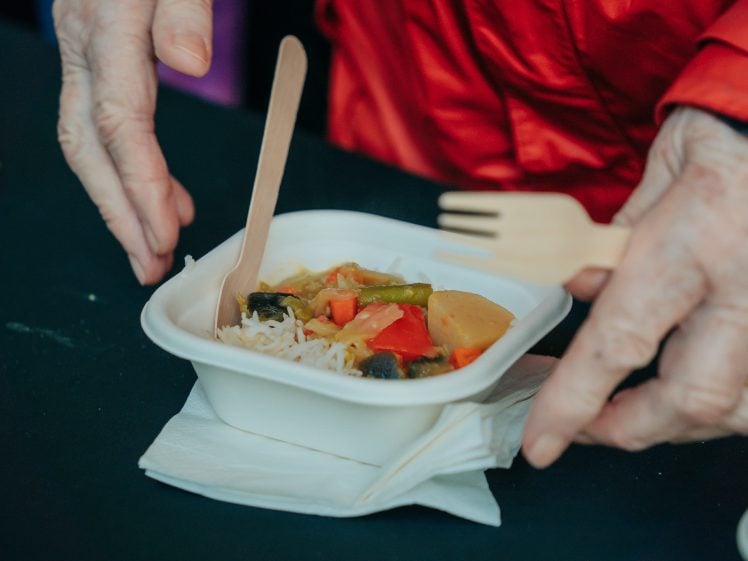 Image of A person holds a compostable container of noodles with vegetables and a wooden fork, over a serviette on a black surface. The person is wearing a red sleeve and a ring.